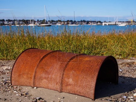 Photo for Closeup photo of rusty barrel half buried in the sand in Coughlin Park in Winthrop outside Boston, Massachusetts, USA, with sunshine, water and sailboats. Winthrop is a town in Suffolk County. - Royalty Free Image