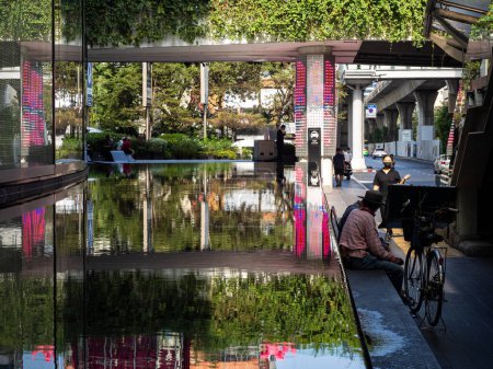 Photo for This photo showcases a reflective pool outside the Emporium Mall, located by Phrom Phong station along Sukhumvit Road in Bangkok, Thailand. In the photo, people wearing face masks can be seen enjoying the outdoor space while maintaining social distan - Royalty Free Image
