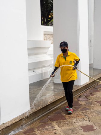 Photo for A person wearing a yellow shirt and a face mask is using a hose to clean the ground at the Wat Pho temple in Bangkok, Thailand. - Royalty Free Image