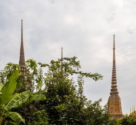 Photo for The golden spires of the Wat Pho temple shine against a cloundy sky, as they emerge from behind the lush greenery that surrounds them in Bangkok, Thailand. - Royalty Free Image