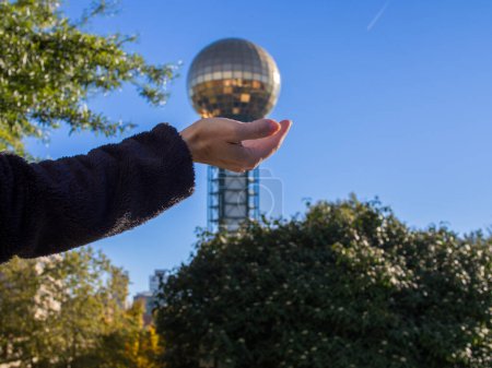 Photo for An enchanting illusion: a female hand holds the golden globe of Sunsphere Tower in Knoxville, Tennessee, USA. Blue sky and lush greenery surround. - Royalty Free Image