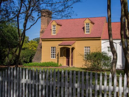 Photo for In Colonial Williamsburg, Virginia, a delightful two-story home features vivid yellow sidings, a bold red roof, and a prominent brick chimney. Against a blue sky, the house stands behind a white fence, radiating classic colonial charm. - Royalty Free Image