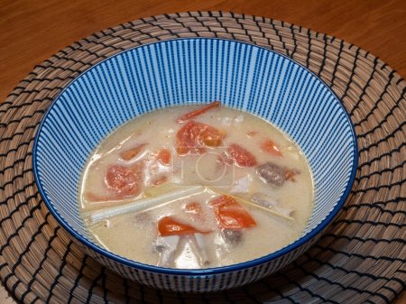 Vegetarian-style Tom Kha Thai soup, featuring coconut milk, tomatoes, mushrooms, and lemongrass, elegantly served in a blue-striped bowl atop a bamboo placemat.