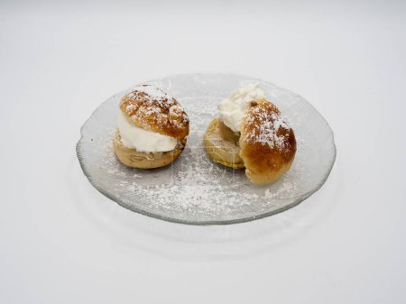 Photo for A side perspective of a classic Swedish semla, also known as fastlagsbulle, adorned with whipped cream, almond paste, and powdered sugar, elegantly presented on a glass plate against a white backdrop. - Royalty Free Image