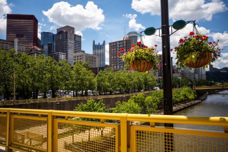 On a sunny day, the yellow Rachel Carson Bridge offers a stunning view of downtown Pittsburgh, with the Allegheny River flowing below, framed by blooming flowers and lush green trees.