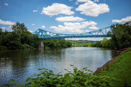 The Boston Bridge spans the Youghiogheny River, connecting to Versailles, Pennsylvania, amidst verdant surroundings under a serene blue sky adorned with fluffy white clouds.