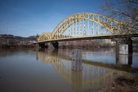 Along the Strip District in Pittsburgh, the yellow Sixteenth Street Bridge casts its reflection upon the tranquil waters of the Allegheny River, framed by a pristine blue sky.