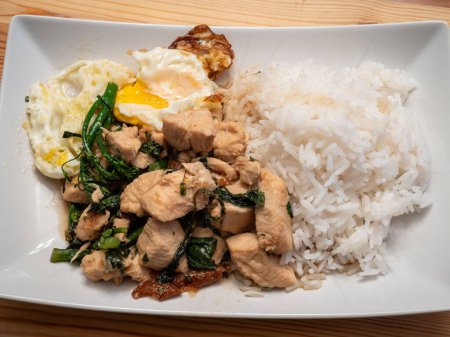 Photo for Indulge in the flavors of Thai basil chicken served with a perfectly fried egg, broccolini, and fragrant jasmine rice, elegantly presented on a white plate atop a rustic wooden board. - Royalty Free Image