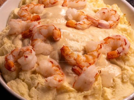 A close-up of a seafood casserole dish, showcasing the rich texture of mashed potatoes contrasting with plump shrimp glistening in a creamy white wine sauce.