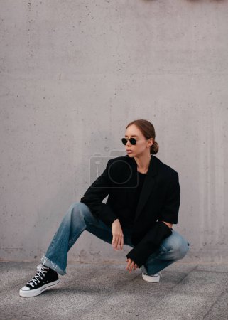 Photo for Model with trending clothes. Urban scene. Youth modeling. Urban trending fashion scene. Low perspective. Street fashion. Young gorgeous woman in black jacket, jeans and sunglasses - Royalty Free Image