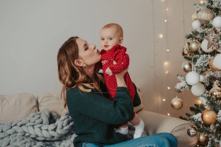 Téléchargez les photos : Mère et bébé en pyjama rouge sourire sur un fond d'arbres de Noël à l'intérieur de la maison - en image libre de droit