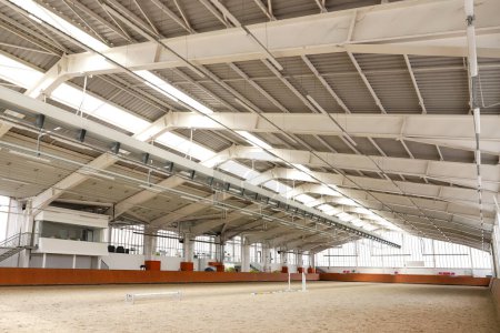 Modern indoor riding arena covering sand for horse trainings. Empty spacious riding hall interior view. Sunlight through windows. Modern equestrian place indoors