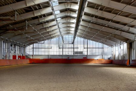 Modern indoor riding arena covering sand for horse trainings. Empty spacious riding hall interior view. Sunlight through windows. Modern equestrian place indoors
