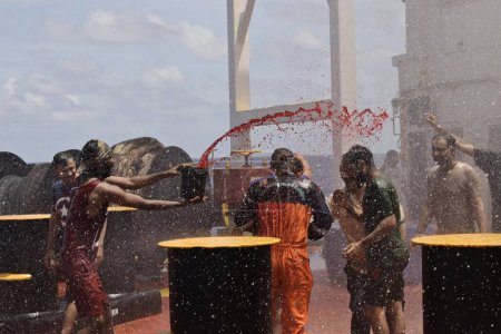 Foto de A bordo del barco, en el mar - 18 de marzo de 2023: marineros indios jugando y celebrando el holi, festival de color bajo la cortina de spray de agua creado para el evento en la cubierta de la caca - Imagen libre de derechos