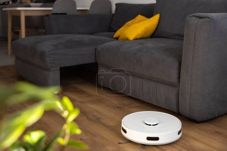 Una aspiradora robot blanca limpia laminado de madera en el suelo en la sala de estar. recoge el polvo cerca del sofá en lugares difíciles y hojas de debajo del sofá grande. sensores infrarrojos y cámaras de control.