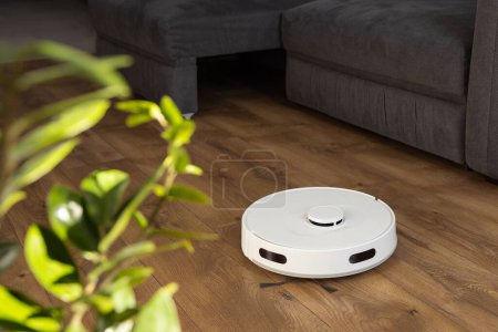 Un aspirateur robot blanc nettoie le stratifié de bois sur le sol dans le salon. recueille la poussière près du canapé dans des endroits difficiles et part de sous le grand canapé. capteurs infrarouges et contrôle des caméras.