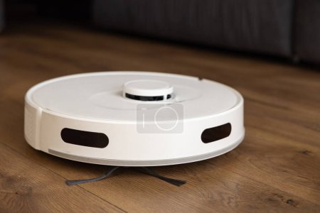 Una aspiradora robot blanca limpia un laminado de madera de roble oscuro en el suelo. recoge polvo cerca del sofá en lugares difíciles. gestión automática de las tecnologías modernas. control de sensores infrarrojos 
