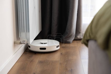 Foto de Una aspiradora robot blanca con un cepillo de barrido, sensores de movimiento infrarrojo y una cámara limpia el suelo cerca de un radiador de calefacción en una habitación. ayuda doméstica - Imagen libre de derechos