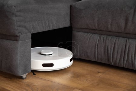 Una aspiradora robot blanca limpia laminado de madera en el suelo. recoge el polvo cerca del sofá en lugares difíciles y hojas de debajo del sofá grande. sensores infrarrojos y control de cámaras. parktronic.