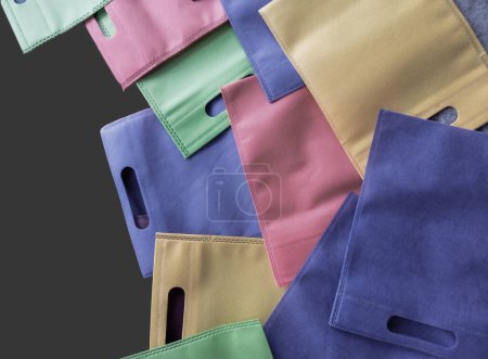 Téléchargez les photos : Collection de sacs fourre-tout aux couleurs pastel isolés sur un fond gris foncé. en polypropylène. pile de sacs non tissés avec poignées perforées - en image libre de droit