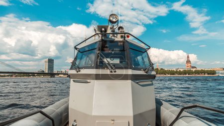 Téléchargez les photos : Bateau de patrouille militaire moderne naviguant sur la rivière avec une ville en arrière-plan et un ciel bleu en toile de fond - en image libre de droit
