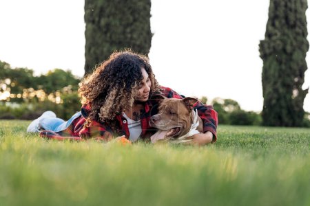 Téléchargez les photos : Jeune femme africaine aux cheveux bouclés couchée dans le parc et embrassant son chien de race. - en image libre de droit