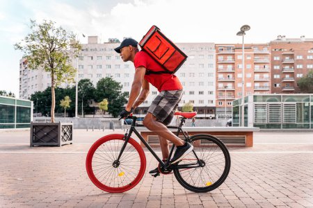 Photo for African delivery man wearing cap riding his bike in the city in his way to deliver a package. - Royalty Free Image