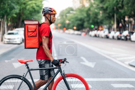 Téléchargez les photos : Un livreur adulte transportant un colis et portant un casque de sécurité traversant la ville avec son vélo. - en image libre de droit