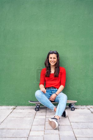 Happy young woman sitting in her long-board against green background and looking at camera.