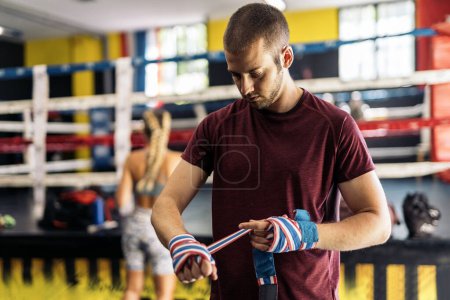 Téléchargez les photos : Homme adulte debout enveloppant les mains avec enveloppement de boxe clored pour l'entraînement dans la salle de gym. Concept de formation. - en image libre de droit