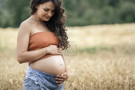 Photo for Pregnant woman standing in wheat field. She's touching and looking her belly. - Royalty Free Image