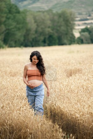 Photo for Pregnant woman walking in wheat field. She's touching her belly and smiling. - Royalty Free Image