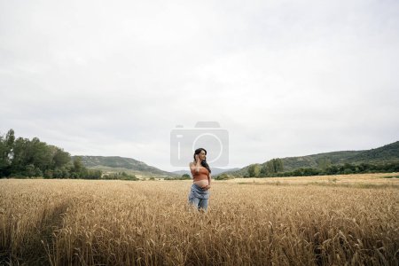 Photo for Pregnant woman standing in wheat field. She's touching her belly and looking to the side. - Royalty Free Image