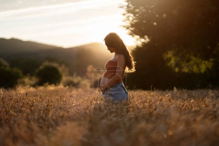 Photo for Side view of pregnant woman standing in wheat field. She's touching and looking her belly. - Royalty Free Image