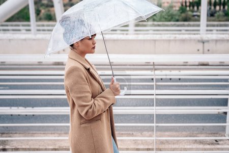 Téléchargez les photos : Jolie femme marchant dans la ville pendant la journée pluvieuse tenant un parapluie transparent. Elle porte du maquillage et regarde devant elle. - en image libre de droit