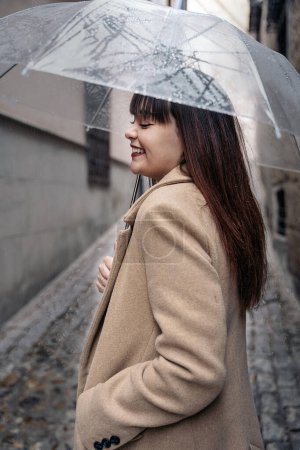 Photo for Young woman holding her umbrella and smiling in the street during rainy day. She is wearing a coat. - Royalty Free Image