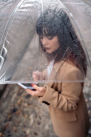 Foto de Mujer joven no reconocida con paraguas usando su teléfono móvil en la calle durante el día lluvioso. - Imagen libre de derechos