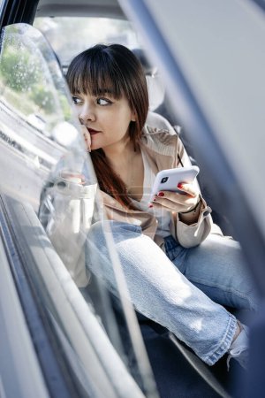 Foto de Mujer feliz sentada en su coche y usando su teléfono móvil. Ella está usando maquillaje y ropa casual. - Imagen libre de derechos