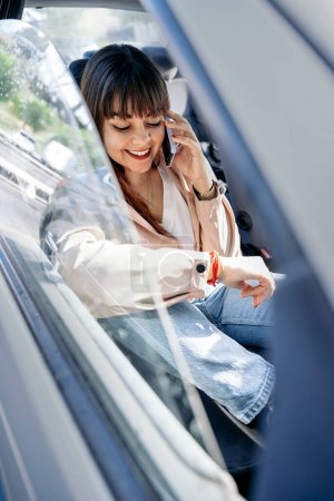 Téléchargez les photos : Femme heureuse assise dans sa voiture et parlant sur son téléphone portable. Elle porte du maquillage et des vêtements décontractés. - en image libre de droit