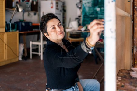 Foto de Artista enfocado de mediana edad pintando en lienzo grande. Ella tiene un estudio de arte en su garaje con luz natural. - Imagen libre de derechos
