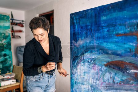 Foto de Pintora femenina feliz relajándose en su pintura de garaje con pintura azul en un lienzo grande. - Imagen libre de derechos