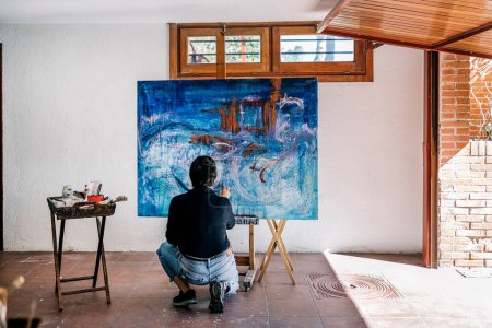 Téléchargez les photos : Peintre féminine méconnue relaxant dans sa peinture de garage avec de la peinture bleue dans une grande toile. - en image libre de droit