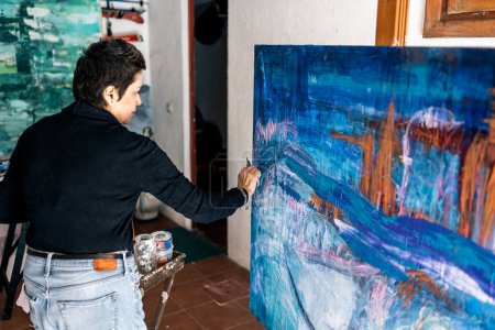 Téléchargez les photos : Peintre féminine focalisée peinture avec des couleurs bleues une œuvre d'art abstrait dans son studio à la maison. - en image libre de droit