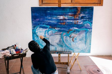 Foto de Pintora femenina no reconocida relajándose en su pintura de garaje con pintura azul en un lienzo grande. - Imagen libre de derechos