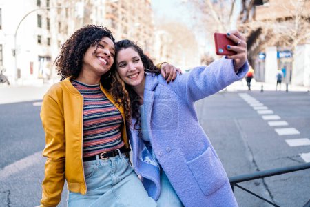 Téléchargez les photos : Belles jeunes filles aux cheveux bouclés souriant et prenant selfie avec téléphone portable dans la rue. - en image libre de droit
