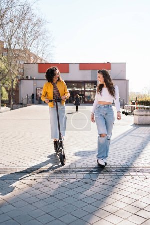 Foto de Chicas de pelo rizado divirtiéndose en la calle usando scooter eléctrico. Están hablando y riendo.. - Imagen libre de derechos