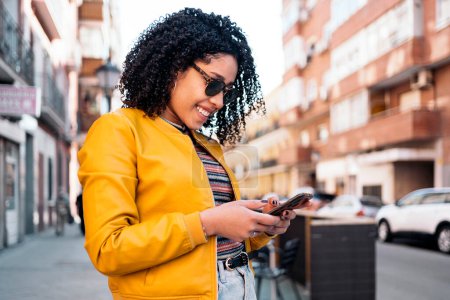 Photo for Beautiful african young woman using her mobile phone in the street and wearing sunglasses. - Royalty Free Image