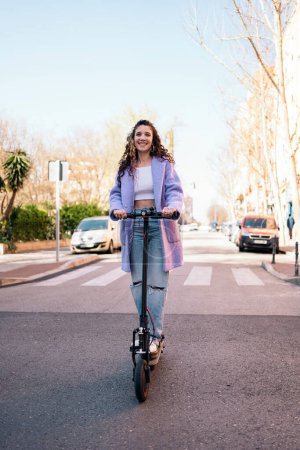 Photo for Cheerful young woman with curly hair using electric scooter in the street and having fun. - Royalty Free Image