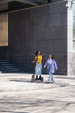 Foto de Chicas guapas divirtiéndose en la calle usando scooter eléctrico. Están hablando y riendo.. - Imagen libre de derechos