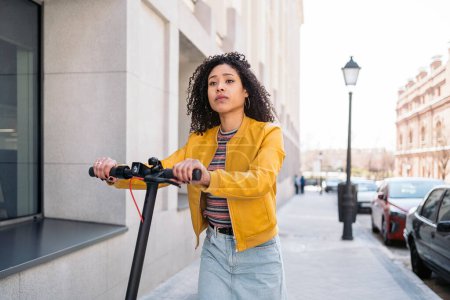 Photo for Young woman with curly hair using electric scooter in the street and looking at front. - Royalty Free Image